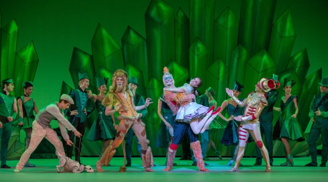 Green emerald background featuring performers from Colorado Ballet dressed as characters from Wizard of Oz - Scarecrow, Tinman, Dorothy