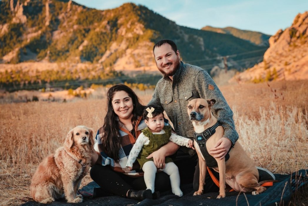 Photo of a family of dad, mom, toddler, and 2 dogs with mountains in the background.