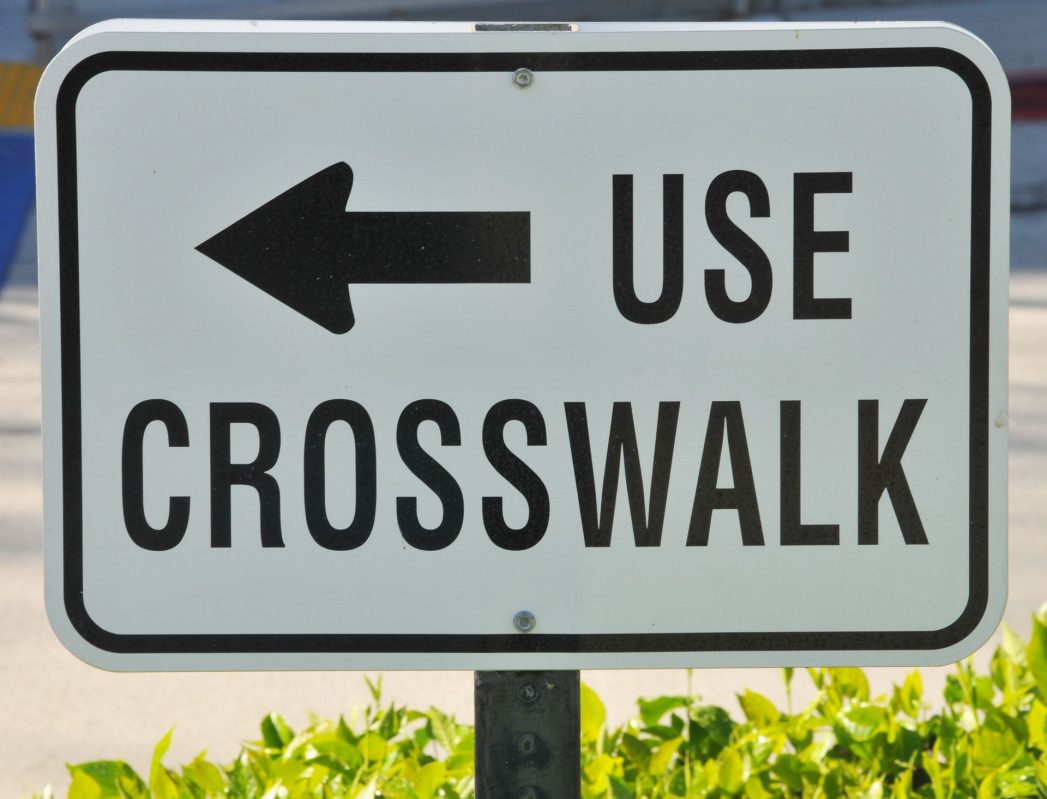 Image of street sign, black lettering on white background, that says "Use Crosswalk."