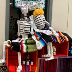 Two larger skeleton figures leaning against each other and sitting on top of the Day of the Dead ofrenda.