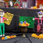 Two larger skeleton figurines standing in front of the Day of the Dead ofrenda.