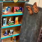 A brown cabinet with the door open to reveal four shelves of Day of the Dead skeleton figurines. A Monarch butterfly is perched on the door.