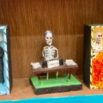 Close up of Day of the Dead skeleton figurines, two are in coffins and one is sitting at a desk typing.